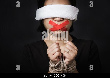 Slave Asian woman blindfold wrapping mouth with red adhesive tape, tied with chains and closed her eyes black background. Freedom speech censorship an Stock Photo
