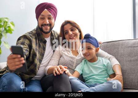 A SIKH MAN TAKING SELFIE ON MOBILE PHONE WITH WIFE AND SON WHILE SITTING TOGETHER ON SOFA Stock Photo