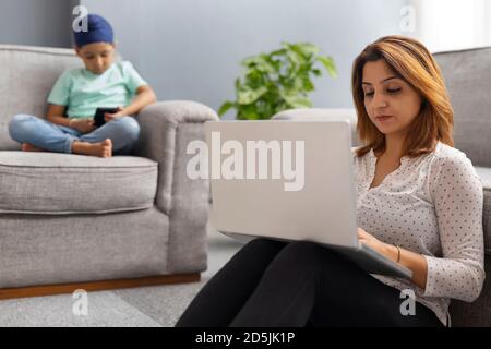 A SIKH SON SITTING ON SOFA WITH  CROSSED LEGS USING MOBILE PHONE WHILE MOTHER WORKS ON LAPTOP SITTING NEARBY Stock Photo