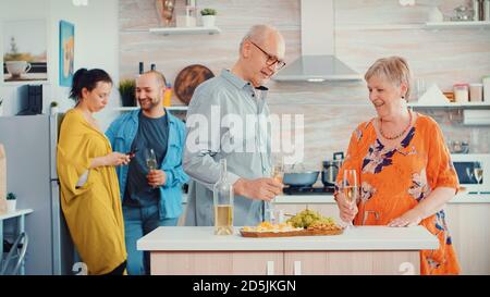 Cheerful old elderly adult family hugging laughing dancing drinking a glass of white wine in front young couple. In the kitchen. Grandma and grandpa embracing smiling and daughter taking a photo Stock Photo