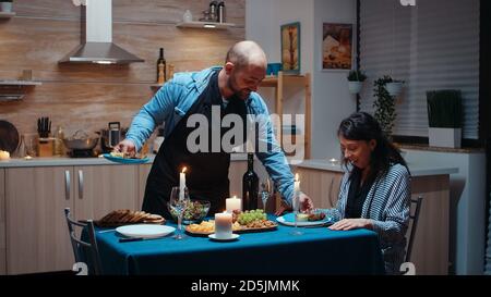 Man preparing festive dinner for his beautiful wife. Young couple talking, sitting at the table in kitchen, enjoying the meal, celebrating their anniversary at home with healty food. Romantic happy Stock Photo