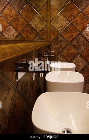 Golden brown design water closet room with bronze color tile on a wall and big mirror three sinks. Stock Photo