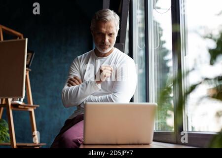 Casual Grey-haired Mature man in eyeglasses working on laptop while sitting in office or cafe near the window Stock Photo