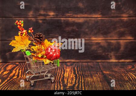 Autumn sale. Shopping cart on wooden background with autumn decor - maple leaves, cones, berries, pumpkin. Stock Photo