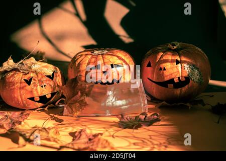 Three halloween pumpkins painted as Jack's lantern, one in protective face mask, fall leaves, magic shadows. New normal. Stock Photo