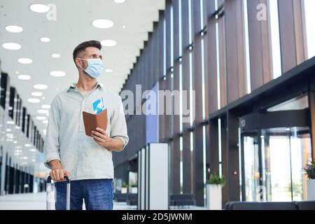 Young man in protective mask with luggage and tickets standing at the airport Stock Photo