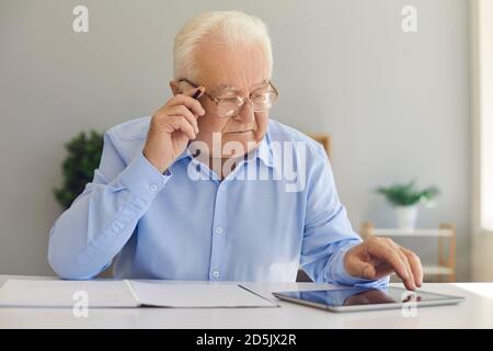 Serious old man sitting at desk at home using tablet computer and taking notes in notebook Stock Photo