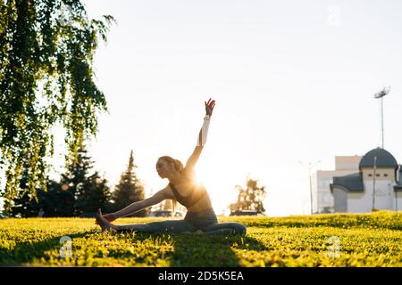 Side view of flexible young woman sitting on yoga mat and making deflections to side, stretching outside Stock Photo