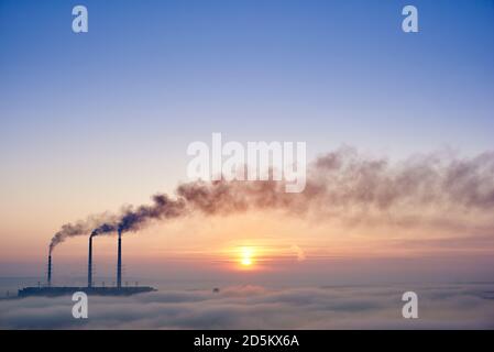 Horizontal snapshot of three smoking stacks of thermal power station on the horizon taken from the hill, pipes are in evening fog on blue sky, copy space Stock Photo