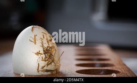 Single isolated straw covered fresh hens egg in wooden holder in kitchen. Dark background ingredients food produce healthy eating. Stock Photo