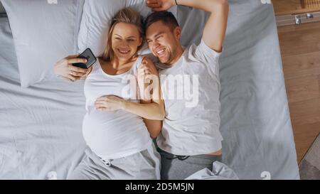 Loving Young Couple Spending Morning in Bed, Pregnant Young Woman Shows Her Partner Something on a Touchscreen Smartphone, Taking Selfie and Sharing Stock Photo