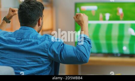 At Home Sports Fan Watches Important Soccer Match on TV, Cheering For His Team, Celebrates Doing YES Gesture after the GOAL Brings Victory to His Team Stock Photo