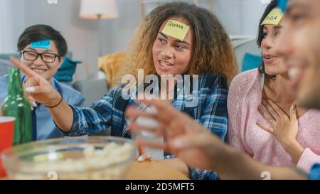 In the Living Room Diverse Group of Fun Loving Friends Playing 'Who am I' game With Sticky Papers Attached to Foreheads. Girls Points Her Finger to Stock Photo