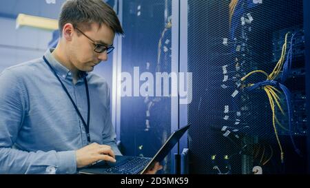 In Data Center IT Engineer Stands Before Working Server Rack Doing Routine Maintenance Check and Diagnostics Using Laptop. Visible Computer Hardware Stock Photo