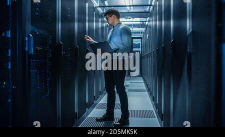 In Data Center IT Engineer Stands Before Working Server Rack Doing Routine Maintenance Check and Diagnostics Using Laptop. Concept of Cloud Computing Stock Photo