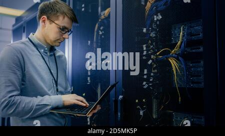 In Data Center IT Engineer Stands Before Working Server Rack Doing Routine Maintenance Check and Diagnostics Using Laptop. Visible Computer Hardware Stock Photo