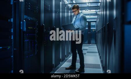 In Data Center IT Engineer Stands Before Working Server Rack Doing Routine Maintenance Check and Diagnostics Using Laptop. Concept of Cloud Computing Stock Photo