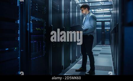In Data Center IT Engineer Stands Before Working Server Rack Doing Routine Maintenance Check and Diagnostics Using Laptop. Concept of Cloud Computing Stock Photo