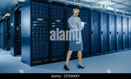 In Data Center: Female IT Specialist Walks along the Row of Operational Server Racks, Uses Laptop to Run Maintenance Programme. Modern High-Tech Stock Photo