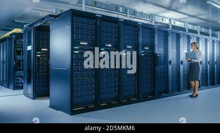 In Data Center: Female IT Specialist Walks along the Row of Operational Server Racks, Uses Laptop to Run Maintenance Programme. Modern High-Tech Stock Photo