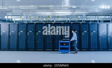In the Modern Data Center: IT Technician Working with Server Racks, Pushes Cart Between Rows of Server Racks. On a Pushcart New Hardware for System Stock Photo