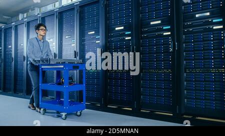 In the Modern Data Center: IT Technician Working with Server Racks, Pushes Cart Between Rows of Server Racks. On a Pushcart New Hardware for System Stock Photo
