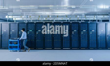 In the Modern Data Center: IT Technician Working with Server Racks, Pushes Cart Between Rows of Server Racks. On a Pushcart New Hardware for System Stock Photo