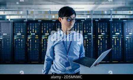 In the Modern Data Center: Portrait of IT Engineer Standing with Server Racks Behind Him, Holding Laptop and Looking at the Camera. Finishing Doing Stock Photo