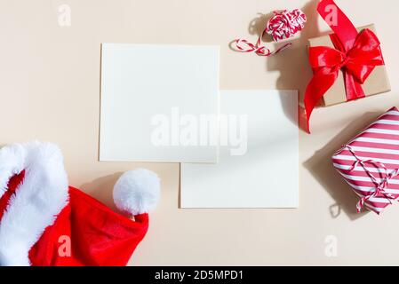 Christmas congratulation mock-up cards with Santa's red hat, festive gift boxes on a light beige background, copy space. Flat lay. Stock Photo