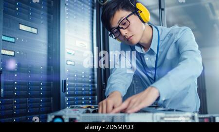 In the Modern Data Center: IT Engineer Wearing Protective Headphones Installs New Hardware for Server Rack. IT Specialist Doing Maintenance, Running Stock Photo