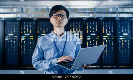 In the Modern Data Center: Portrait of IT Engineer Standing with Server Racks Behind Him, Holding Laptop and Looking at the Camera. Finishing Doing Stock Photo