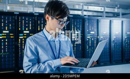 In the Modern Data Center: Portrait of IT Engineer Standing with Server Racks Behind Him, Doing Maintenance and Diagnostics Procedure Using Laptop. Stock Photo