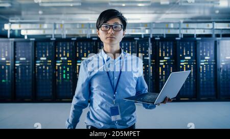 In the Modern Data Center: Portrait of IT Engineer Standing with Server Racks Behind Him, Holding Laptop and Looking at the Camera. Finishing Doing Stock Photo