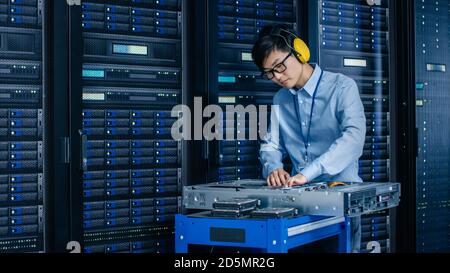 In Data Center: IT Engineer Wearing Protective Muffs Installs New Hardware for Server Rack. Specialist Doing Maintenance, Running Diagnostics and Stock Photo