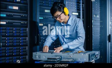In the Modern Data Center: IT Engineer Wearing Protective Muffs Installs New Hardware for Server Rack. IT Specialist Doing Maintenance, Updating Stock Photo
