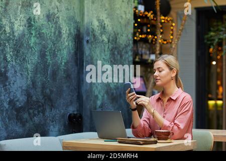 Young businesswoman using mobile phone while sitting at the table with laptop and working at cafe Stock Photo