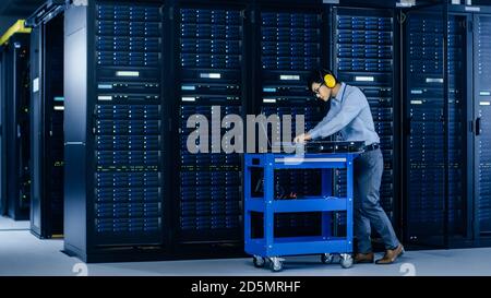 In Data Center: IT Engineer Wearing Protective Muffs Installs New Hardware for Server Rack. Specialist Doing Maintenance, Running Diagnostics and Stock Photo