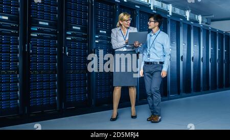 In the Modern Data Center: Engineer and IT Specialist Work with Server Racks, Talking, Doing System Maintenance Check with Laptop Computer Stock Photo