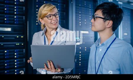 In the Modern Data Center: Engineer and IT Specialist Walking alongside Server Racks, Talking, Doing System Maintenance Check with Laptop Computer Stock Photo