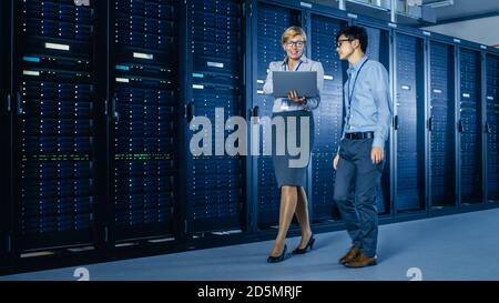 In the Modern Data Center: Engineer and IT Specialist Work with Server Racks, Talking, Doing System Maintenance Check with Laptop Computer Stock Photo