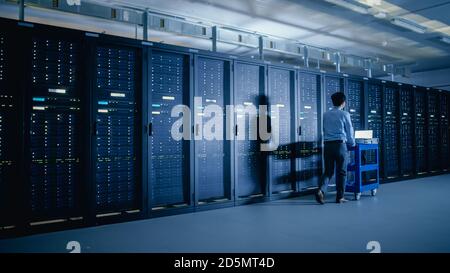 In the Modern Data Center: IT Engineer Doing Maintenance and Diagnostics. Pushes Cart alongside Server Racks. Planned System Equipment Update and Stock Photo