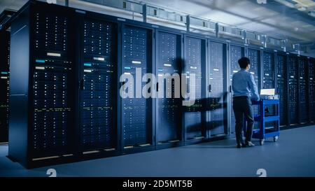 In the Modern Data Center: IT Engineer Doing Maintenance and Diagnostics. Pushes Cart alongside Server Racks. Planned System Equipment Update and Stock Photo