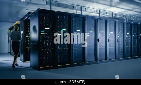 In Data Center: Female IT Technician Walks Alongside Row of Server Racks, Uses Laptop Computer to Run Maintenance Diagnostics so that Mainframe Works Stock Photo