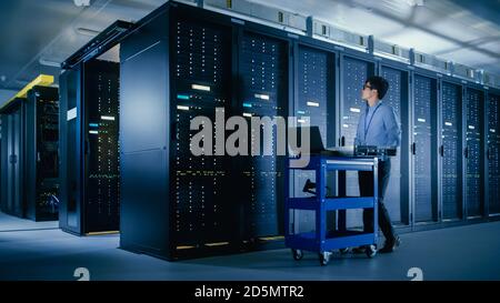 In the Modern Data Center: IT Engineer Doing Maintenance and Diagnostics. Pushes Cart alongside Server Racks, Ready to Install New Hardware for Stock Photo