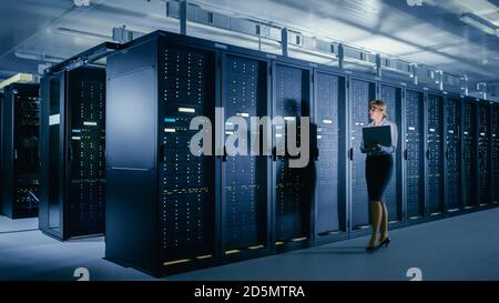 In Data Center: Female IT Technician Walks Alongside Row of Server Racks, Uses Laptop Computer to Run Maintenance Diagnostics so that Mainframe Works Stock Photo