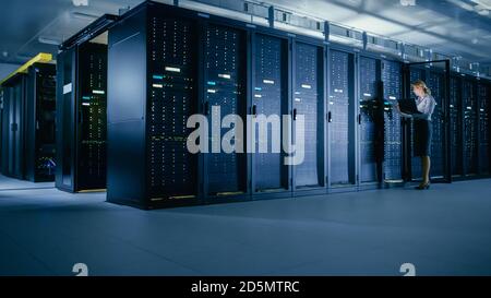 In Data Center: Female IT Technician Stands before Open Server Rack Cabinet, Uses Laptop Computer to Run Maintenance Diagnostics so that Mainframe Stock Photo