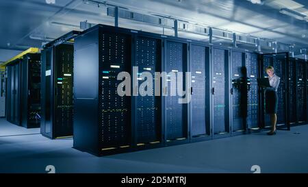 In Data Center: Female IT Technician Stands before Open Server Rack Cabinet, Uses Laptop Computer to Run Maintenance Diagnostics so that Mainframe Stock Photo