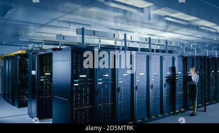 In Data Center: Female IT Technician Stands before Open Server Rack Cabinet, Uses Laptop Computer to Run Maintenance Diagnostics so that Mainframe Stock Photo