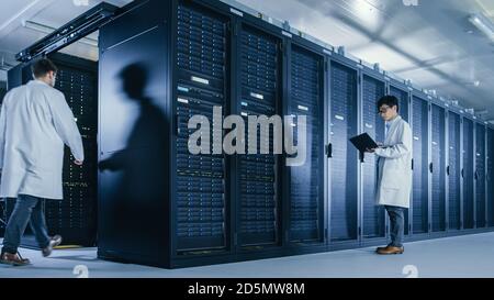In Data Center: Male IT Technician Wearing White Coat Stands Beside Server Rack Cabinet, Uses Laptop Computer to Run Maintenance Diagnostics Tools Stock Photo