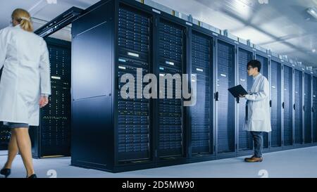 In Data Center: Male IT Technician Wearing White Coat Stands Beside Server Rack Cabinet, Uses Laptop Computer to Run Maintenance Diagnostics Tools Stock Photo